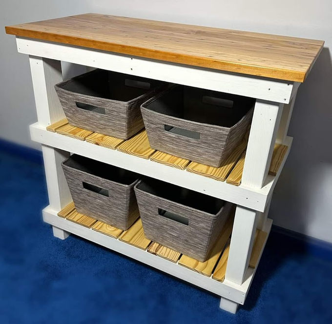 A white two-tier storage island with a warm butcher-block top and four woven baskets on slatted shelves.